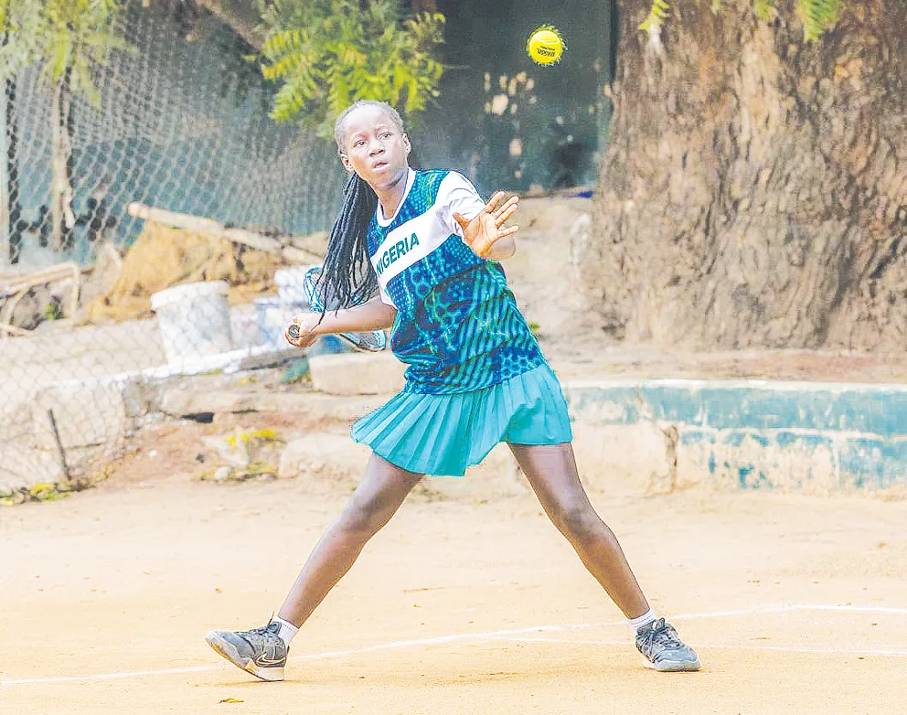 Young Nigerian tennis player reaching toward a ball during a match on an outdoor court