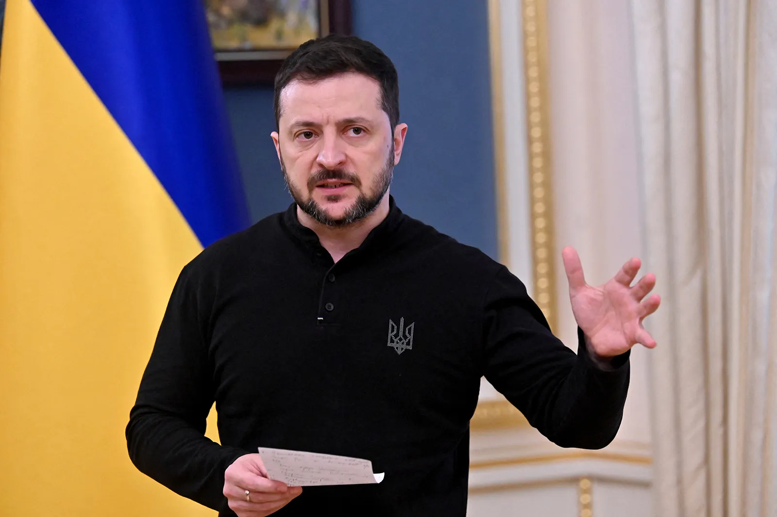 Ukrainian President Volodymyr Zelensky speaking indoors with a Ukrainian flag behind him