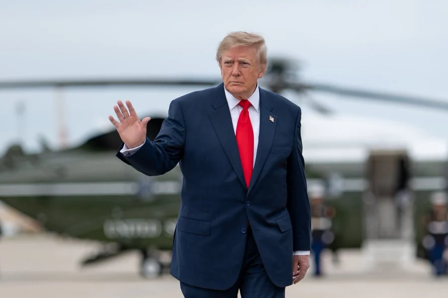 Donald Trump wearing a dark suit and red tie while raising one hand near an aircraft