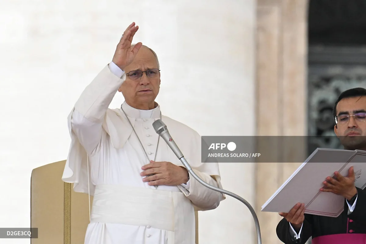 Pope Leo XIV in white papal attire raising one hand while standing at a microphone